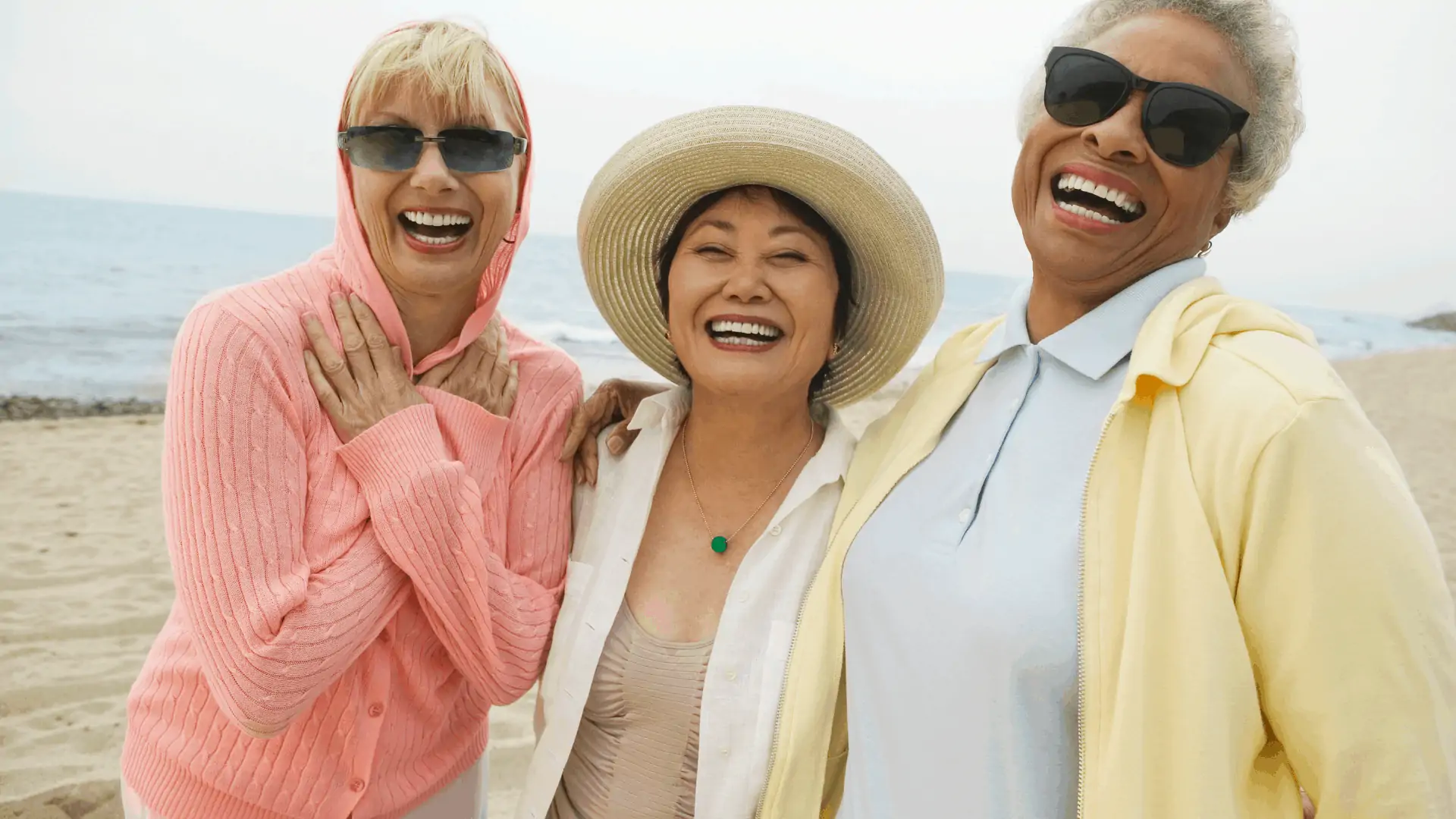 Three female friends who are older smile and laugh for the camera on the beach. 